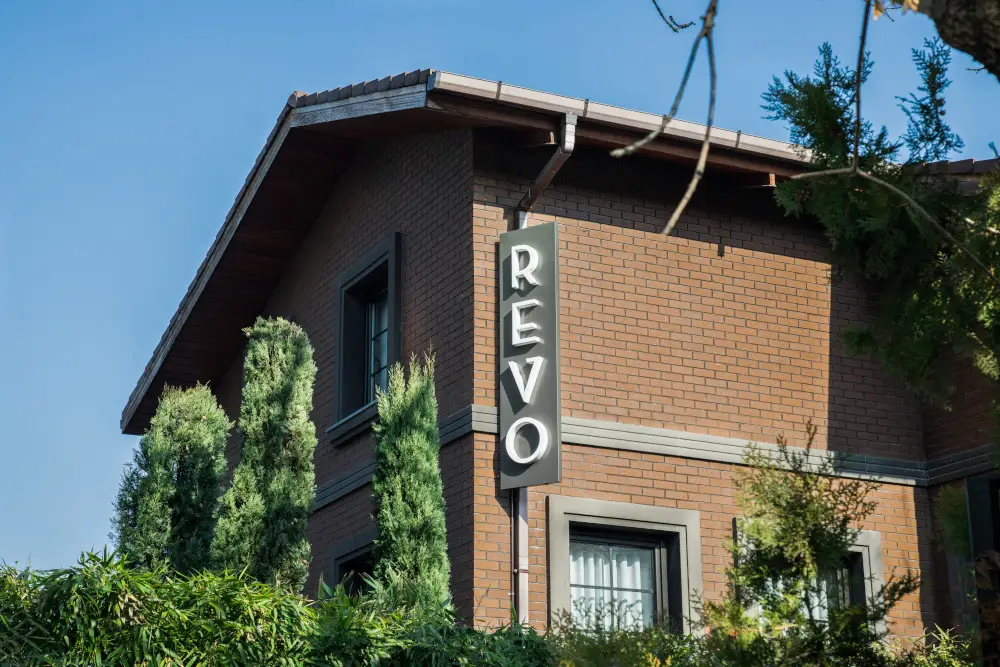 Modern brick building with "REVO" sign, surrounded by lush greenery and clear blue sky.